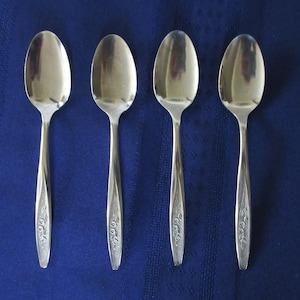 May include: Four silver spoons with ornate designs on the handles. The spoons are arranged in a row on a blue surface.