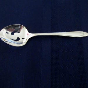 Może przedstawiać: Srebrna łyżka szczelinowa z polerowanym wykończeniem. Łyżka ma owalną miseczkę z dekoracyjnymi wycięciami i długim, smukłym uchwytem. Łyżka jest przeznaczona do serwowania i odcedzania potraw. Tło to ciemnoniebieska tkanina.