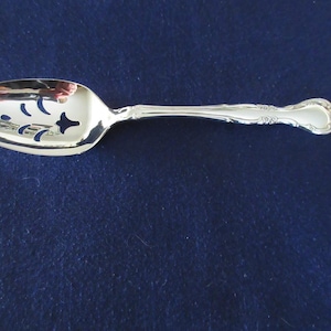 May include: A silver-colored serving spoon with a decorative handle and a slotted bowl. The bowl has a series of cut-out designs. The spoon is set against a dark blue background.