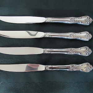 Può includere: Quattro coltelli da tavola d'argento con manici decorati. I coltelli hanno una finitura lucida e sono disposti in fila su una superficie verde scuro.