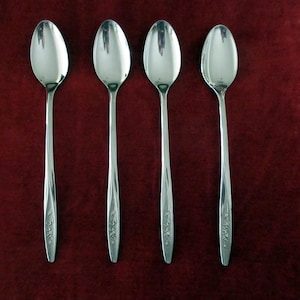 May include: Four polished silver spoons displayed on a deep red surface. Each spoon features a long, slender handle with a floral pattern near the base. The oval-shaped spoon bowls reflect the light, creating a bright contrast.