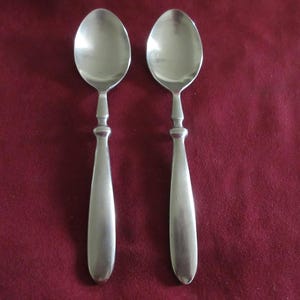 May include: Two matching silver-toned metal spoons with smooth, rounded bowls and straight, tapered handles. Each spoon features a distinct decorative collar detail where the handle meets the bowl, providing a clean and uniform appearance.