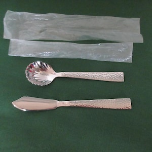 Könnte beinhalten: Zwei silberne Servierutensilien: ein Löffel mit einer gewellten Schale und ein Buttermesser, beide mit strukturierten Griffen. Die Utensilien werden auf einer grünen Oberfläche mit zwei transparenten Kunststoffverpackungen darüber präsentiert.
