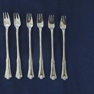 May include: A set of six silver-toned metal cocktail forks arranged in a vertical row. Each fork features a slender handle with a flared base and a decorative shell-like motif at the bottom, ending in three short, pointed tines at the top.