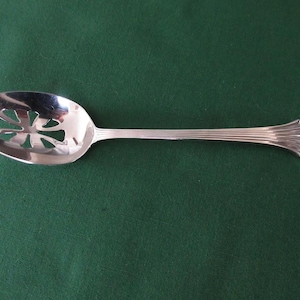 Puede incluir: Una cuchara de servir plateada con un cuenco ranurado y un mango decorativo. El cuenco tiene un diseño calado en forma de flor. El mango es acanalado y termina en un diseño en forma de abanico. La cuchara está sobre una superficie verde oscuro.