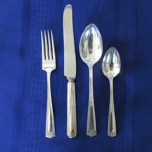 May include: A set of four silver-colored flatware pieces: a fork, a knife, a soup spoon, and a teaspoon. The utensils are arranged on a blue tablecloth. The knife has a decorative handle.