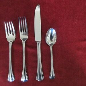 May include: A set of four stainless steel silverware pieces: two forks, a knife, and a spoon. The silverware has a shiny finish and a decorative, fluted design on the handles. The background is a solid, dark red color.