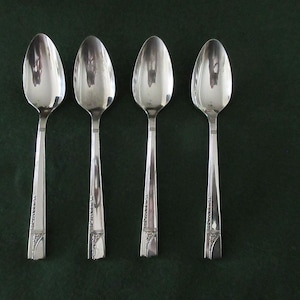 May include: Four silver spoons arranged vertically. Each spoon has a rounded bowl and a long, slender handle with a decorative design near the base. The spoons are reflective, with the surrounding environment mirrored on their surfaces.