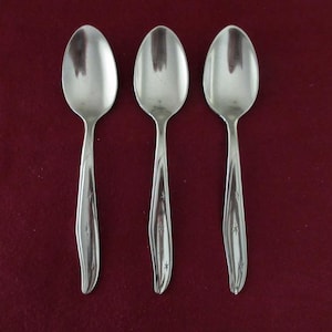 May include: Three silver spoons displayed on a dark red surface. Each spoon has a rounded bowl and a handle with a star pattern. The spoons are made of a shiny metal, reflecting the light. The spoons are arranged vertically.