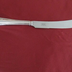 May include: A silver butter knife with a decorative handle and a serrated edge. The knife is made of stainless steel and is displayed on a red surface. The knife has a curved blade and a pointed tip.