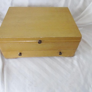 May include: A light-colored wooden box with a hinged lid and a drawer. The box has a rectangular shape with rounded edges and small, dark knobs. The wood grain is visible, and the box appears to be vintage.