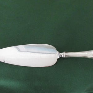 May include: A silver cake server with a wide, oval blade and a decorative handle. The handle features an oval design near the end. The server is set against a dark green background.