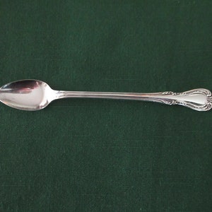 May include: A silver spoon with an ornate handle and a rounded, oval-shaped bowl. The spoon is lying on a dark green surface.