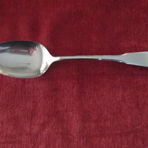 May include: A silver spoon with a rounded bowl and a long, slender handle. The spoon is lying on a red surface.
