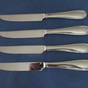 May include: A set of four silver-toned metal dinner knives arranged horizontally. Each knife features a smooth, flat blade and a handle with a decorative beaded edge and a rounded, shell-like motif at the very end of the handle grip.