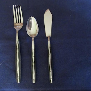 Könnte beinhalten: Ein Besteckset aus Edelstahl mit Gabel, Löffel und Buttermesser. Die schwarzen Griffe sind mit einem Bambusmuster versehen. Die Artikel sind auf einer dunkelblauen Oberfläche angeordnet.