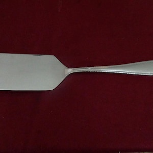May include: A silver-toned metal cake server with a flat, rectangular blade and a slender, rounded handle. The handle features a subtle beaded or textured edge detail that extends from the base of the blade to the end of the handle.
