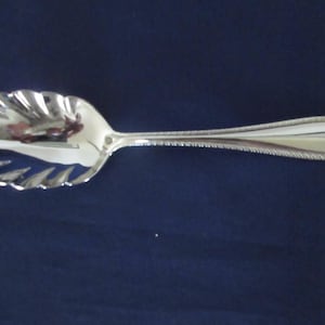 May include: A polished silver-toned serving spoon featuring a bowl shaped like a scalloped shell. The handle has a textured beaded edge detail that extends along the sides, ending in a rounded tip with a decorative embossed pattern.