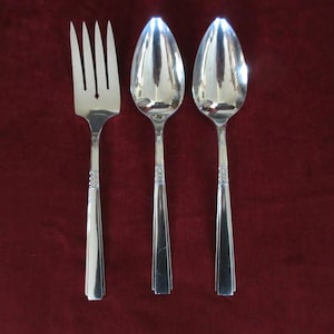 May include: A silver fork and two spoons are arranged on a burgundy background. The silverware has a simple, elegant design with a decorative pattern on the handles. The spoons have a classic oval bowl shape.
