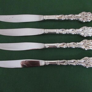 May include: Four silver butter knives with ornate, detailed handles and curved blades are arranged horizontally against a dark green background. The knives are reflective, showing the surrounding environment. The handles have a floral design.
