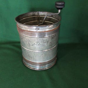 May include: Vintage metal food mill with a hand crank and wooden handle. The cylindrical mill has horizontal bands and the name "Androck" is printed on the side. The mill is made of silver-colored metal with some rust-colored accents.