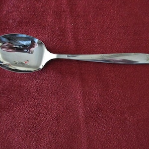 Könnte beinhalten: Ein glänzender, silberner Löffel mit einer abgerundeten Schüssel und einem schlanken Griff. Der Griff hat ein dekoratives Sternmuster am Ende. Der Löffel liegt auf einem tiefroten Hintergrund.