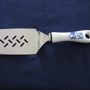 Op de afbeelding: Een roestvrijstalen spatel met sleuven en een witte handgreep met blauwe bloemmotieven. De handgreep heeft een gat om op te hangen. De spatelkop heeft een reeks diagonale sleuven.