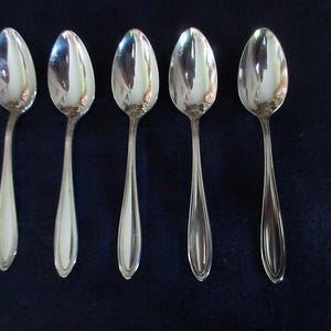 May include: Five silver spoons arranged in a row. Each spoon has a rounded bowl and a long, slender handle. The spoons are reflective, with the surrounding environment mirrored on their surfaces. The spoons are set against a dark blue background.
