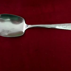 May include: A polished silver serving spoon with a large, oval bowl and a long handle. The handle has a decorative chevron pattern near the end. The spoon is displayed against a deep red background, accentuating its reflective surface.