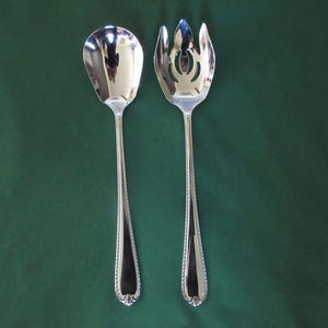 May include: A pair of polished silver-tone metal salad serving utensils consisting of a solid rounded spoon and a matching three-pronged fork. Both pieces feature a slender handle with a decorative beaded edge and a smooth, reflective finish.