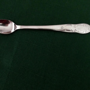 May include: A silver spoon with a rounded bowl and a long, slender handle. The handle features decorative etched patterns near the end. The spoon is set against a dark green background, creating a contrast that highlights its polished surface.
