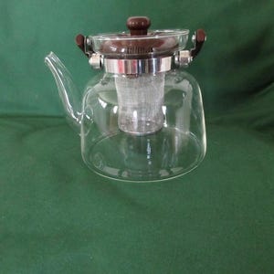 May include: A clear glass teapot featuring a curved spout and a brown plastic lid with a matching handle. The teapot includes a removable stainless steel mesh infuser basket suspended inside the main glass body, secured by a metal band.