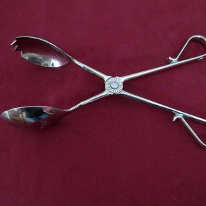 Puede incluir: Pinzas de servir plateadas con extremos en forma de cuchara y un diseño floral decorativo en el punto de pivote. Las pinzas tienen asas de bucle. El utensilio para servir se muestra sobre un fondo burdeos.