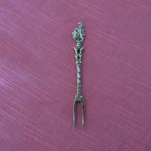 May include: An ornate, silver-toned serving fork with a decorative handle and two tines. The handle features intricate floral and scrollwork designs, with a twisted central section. The fork is set against a pink background.