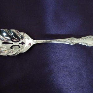 May include: A silver serving spoon with a decorative handle and a scalloped bowl. The bowl has a pierced design. The spoon is set against a dark purple background.