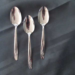 May include: Three polished silver spoons with floral handle detailing are displayed on a dark grey, striped surface. The spoons reflect the surroundings. The spoons vary in size, indicating a dining set. The spoons are made of stainless steel.