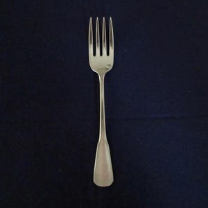 May include: A silver fork with four tines, set against a dark blue background. The fork has a long, slender handle that widens slightly at the base. The tines are evenly spaced and pointed.