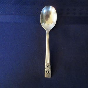 Puede incluir: Una cuchara plateada con un cuenco ovalado y un mango decorativo. El mango presenta líneas verticales y un diseño ornamentado cerca de la base. La cuchara está sobre un fondo azul oscuro.