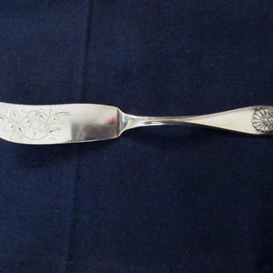 May include: A silver butter knife with a curved blade and a decorative floral design. The handle features a detailed floral pattern at the end. The knife is displayed on a dark blue background.