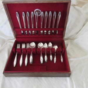 May include: A wooden silverware chest with a red velvet interior, showcasing a complete set of polished silver flatware. The set includes forks, knives, and spoons, neatly arranged for storage and display. The chest is open, revealing the contents.