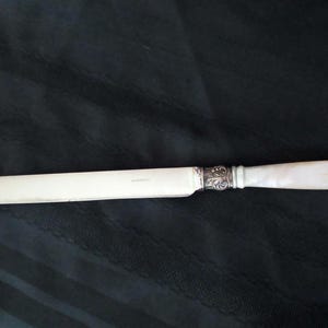 May include: A butter knife with a silver blade and a decorative handle. The handle is white with a floral design at the base and a carved end. The knife is set against a dark background.