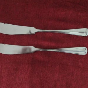 Könnte beinhalten: Zwei silberne Buttermesser mit abgerundeten Griffen und leicht gebogenen Klingen.