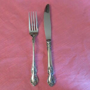 May include: A silver fork and knife set with ornate detailing. The fork has four tines, and the knife has a serrated edge. Both utensils have decorative handles. The set is displayed on a pink tablecloth.