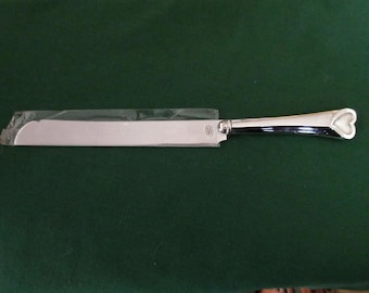 May include: A silver cake knife with a serrated blade and a heart-shaped handle. The handle has a black accent and is wrapped in plastic. The knife is on a dark green surface.