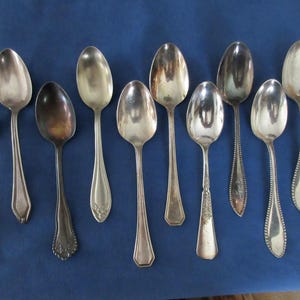 Puede incluir: Una colección de diez cucharas de plata antiguas de varios diseños, dispuestas sobre una superficie azul. Las cucharas varían en tamaño y forma, algunas con detalles ornamentados en el mango y otras con formas más simples y elegantes. Las cucharas son reflectantes.