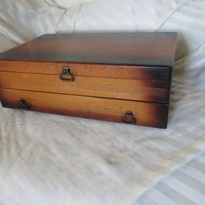 May include: A wooden box with a hinged lid and metal hardware. The box is stained in shades of brown, with a darker top and lighter sides. The box has a rectangular shape and is likely used for storage.