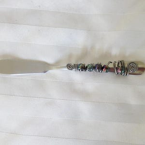 May include: A silver butter knife with a decorative handle. The handle is adorned with wire wrapping and colorful beads. The knife has a flat, wide blade and a spiral design at the end of the handle.