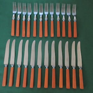 Puede incluir: Un juego de veintitrés piezas de cubiertos de acero inoxidable que consta de once tenedores y doce cuchillos. Cada pieza presenta un mango rectangular de color naranja sólido y una cabeza de metal pulido. Los artículos están dispuestos en dos filas.