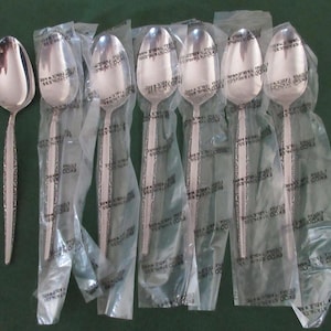 May include: A collection of stainless steel spoons, some individually wrapped in clear plastic. The spoons have a decorative pattern on the handles and a polished, reflective surface. The spoons are arranged on a dark green surface.