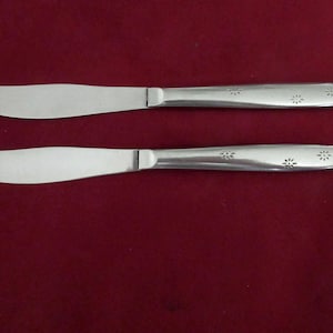 May include: Two stainless steel butter knives with a floral design on the handle. The blades are curved and the handles have a series of small, engraved flower shapes. The knives are set against a dark red background.